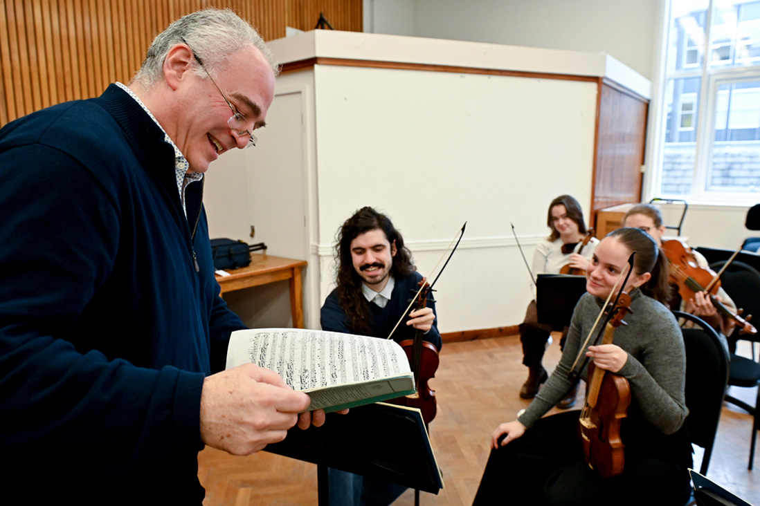 A male professor looking at music sheets talking to students holding their violins and chatting.